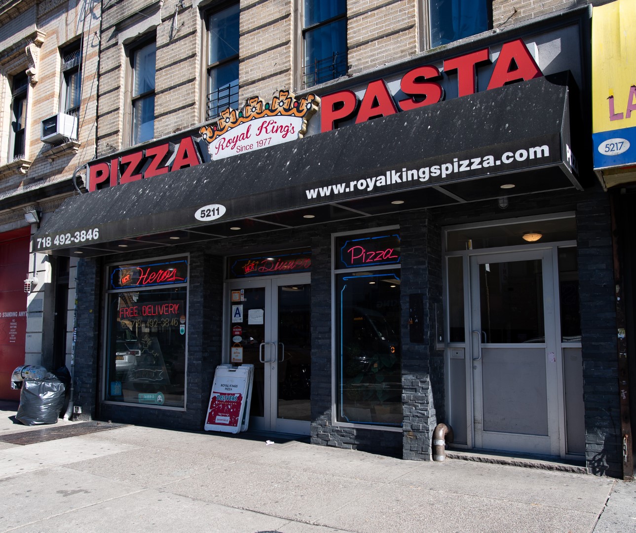 Exterior of a pizza and pasta restaurant named "royal kings pizza" featuring a black awning with red and yellow lettering, displaying advertisements for pizza and free delivery.