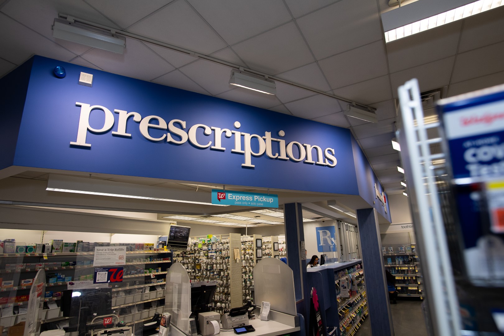 Blue "prescriptions" sign above a pharmacy counter inside a retail store, indicating where customers can pick up their medications.