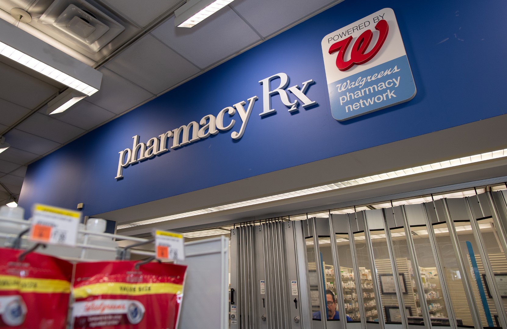 Interior of a pharmacy with "pharmacy rx" sign, featuring the walgreens logo, above the medication counter partially visible behind a display of products.