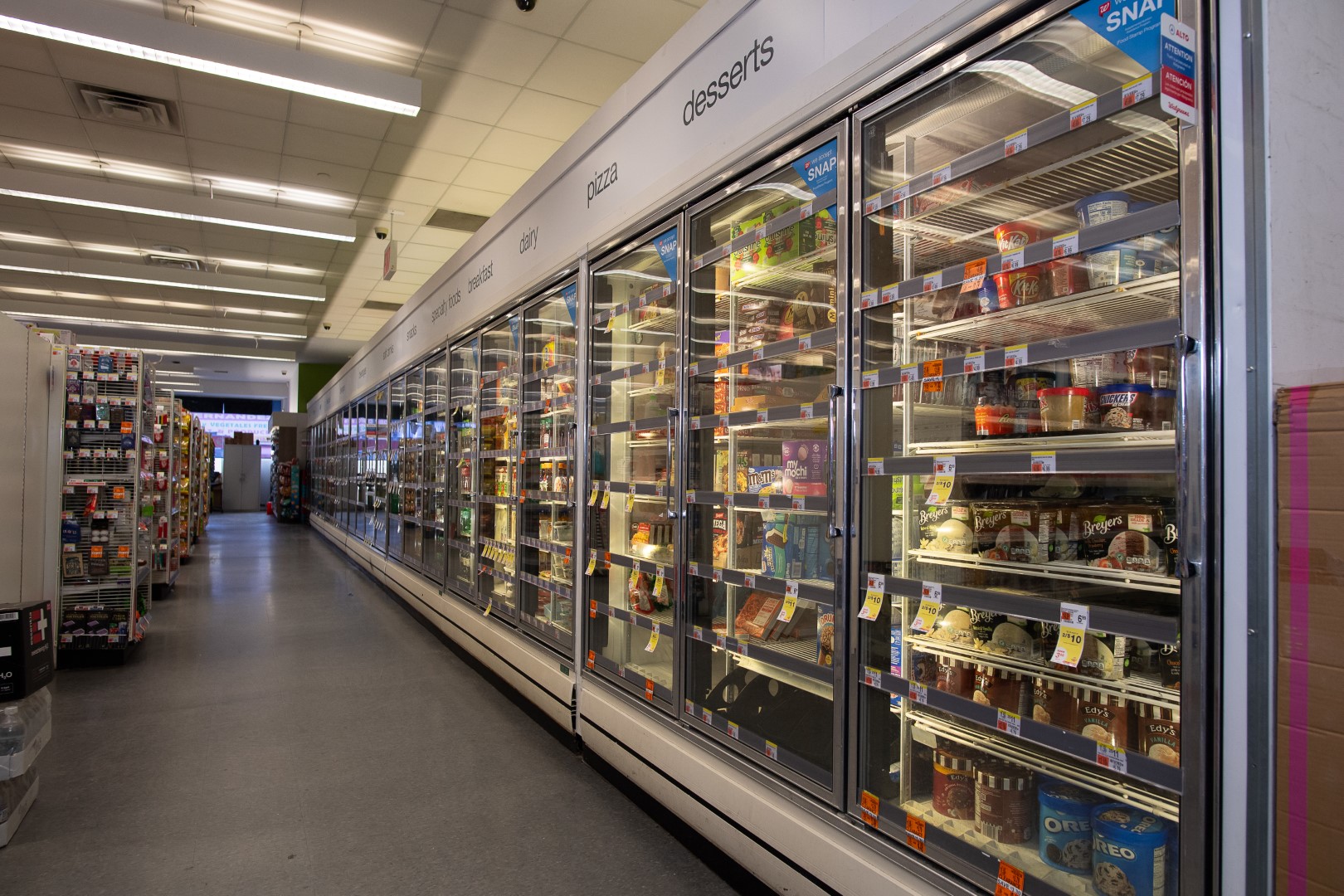 A brightly lit supermarket aisle with a row of refrigerated shelves filled with an assortment of desserts and frozen foods.