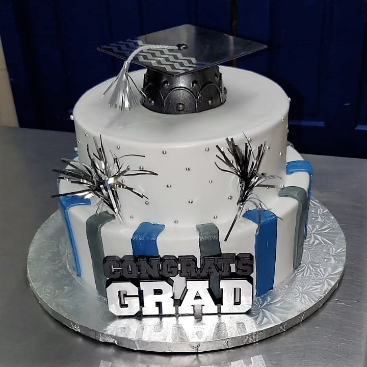 A graduation-themed cake with a two-tier design, decorated in white and silver. the top features a black graduation cap, and the base has the message "congrats grad" with diploma accents.