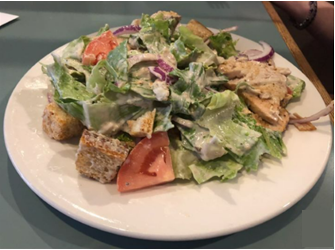 A plate of caesar salad with chunks of grilled chicken, fresh lettuce, tomato wedges, croutons, red onions, and creamy dressing on a table.