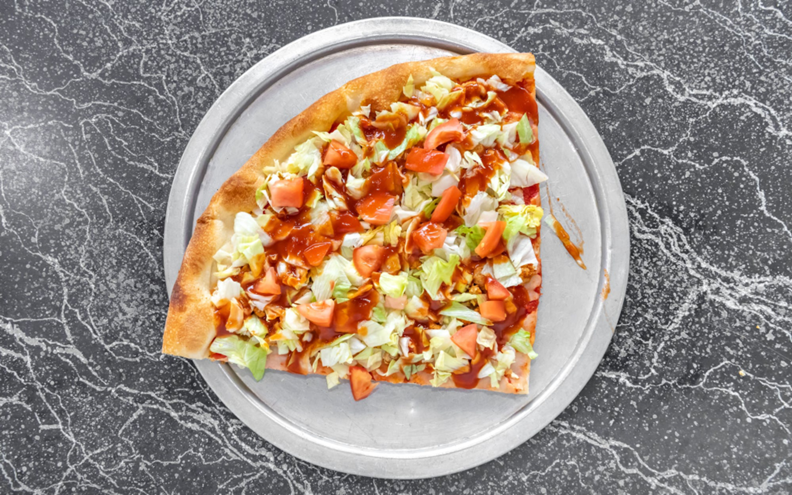 A slice of pizza topped with diced tomatoes and shredded lettuce on a metal tray, placed on a mottled gray surface.