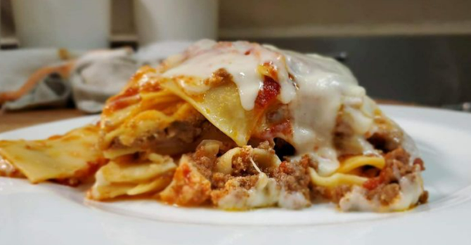 A slice of lasagna on a white plate, showing layers of pasta, meat sauce, and melted cheese, with a blurred kitchen background.