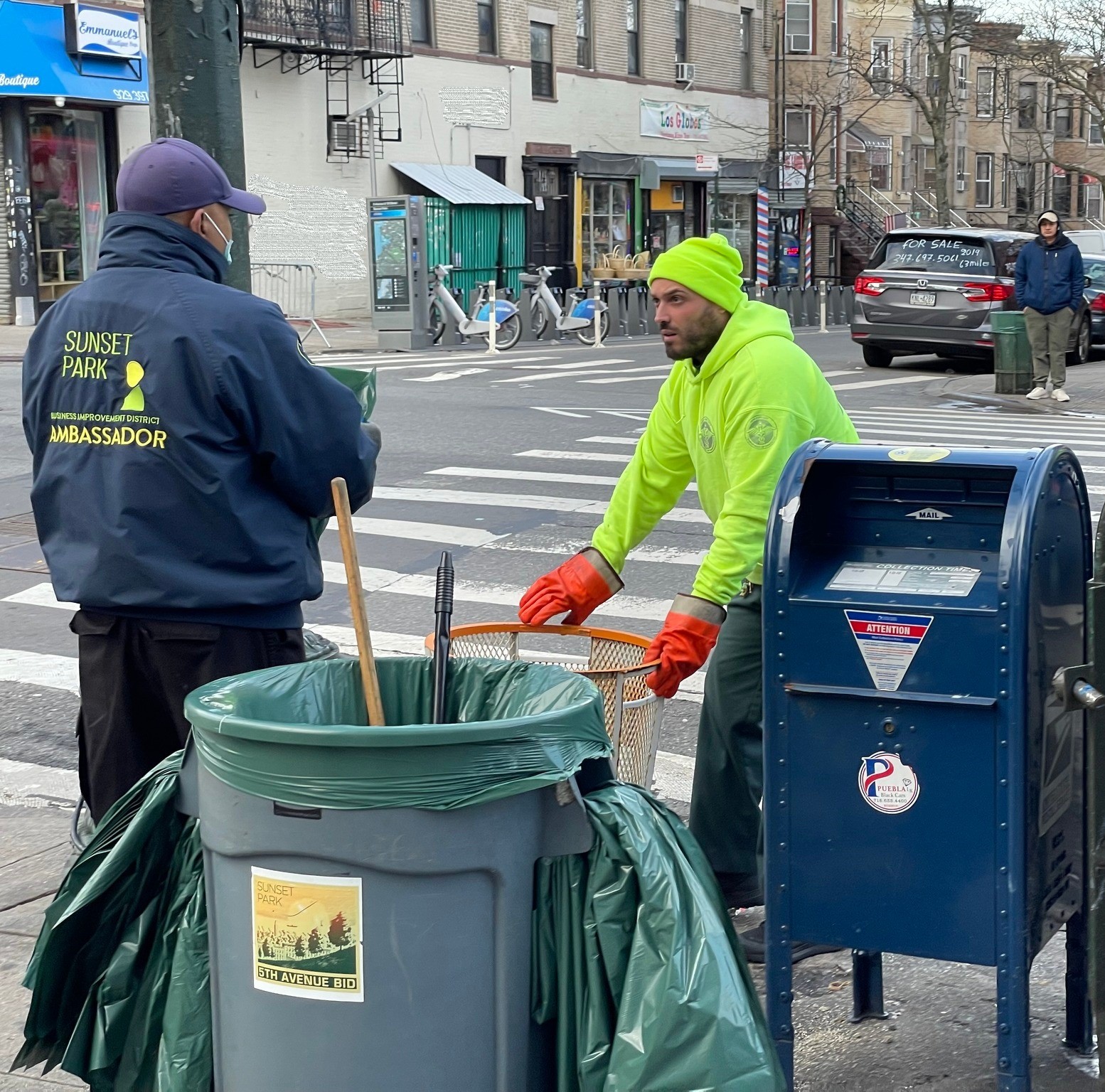 Two sanitation workers in brightly colored jackets chat near a trash bin and mailbox on a busy city street. one worker is handling a broom and dustpan.