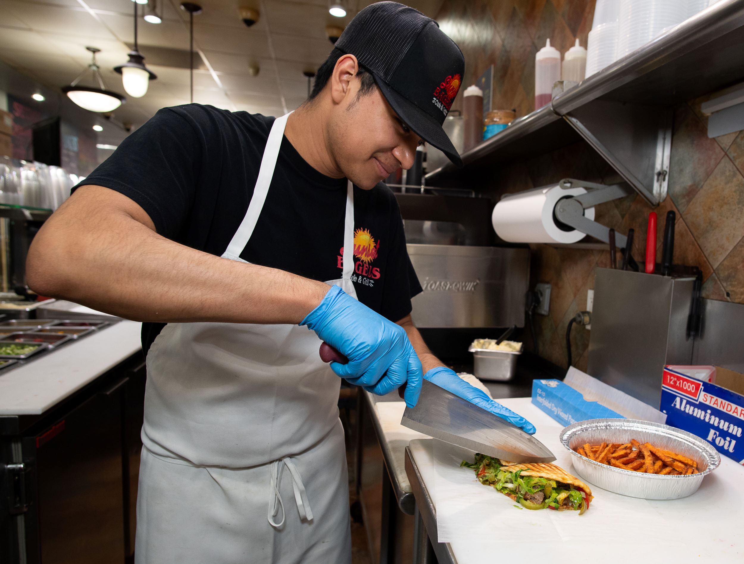 A man wearing a cap, apron, and gloves prepares food in a kitchen, smiling as he slices a wrap with a knife, with a bowl of salad and sweet potato fries nearby.
