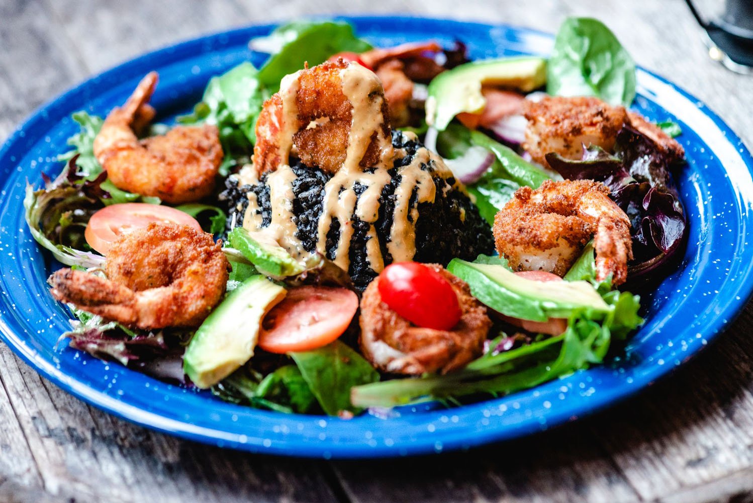 A vibrant shrimp salad with avocado, tomatoes, mixed greens, and a drizzle of creamy dressing, served on a blue-rimmed plate on a wooden table.