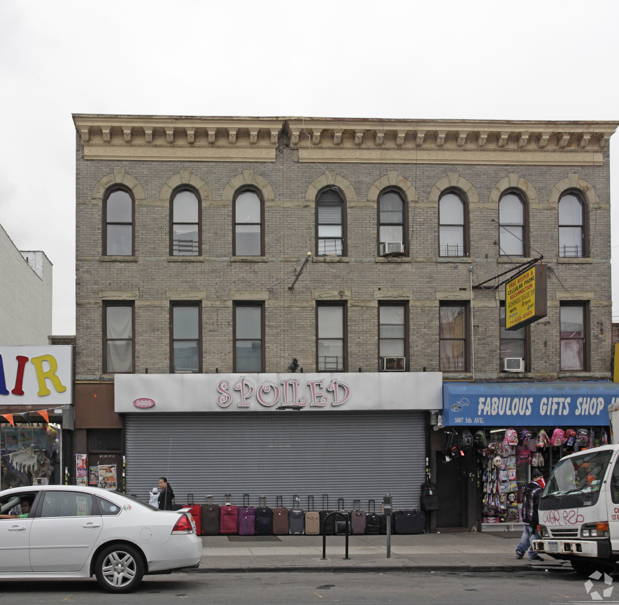 A street view featuring a three-story brick building with arched windows. The first floor has businesses, including a store called "SPOILED" with a closed shutter and sidewalk display of suitcases, and a shop named "FABULOUS GIFTS SHOP" with various items outside.