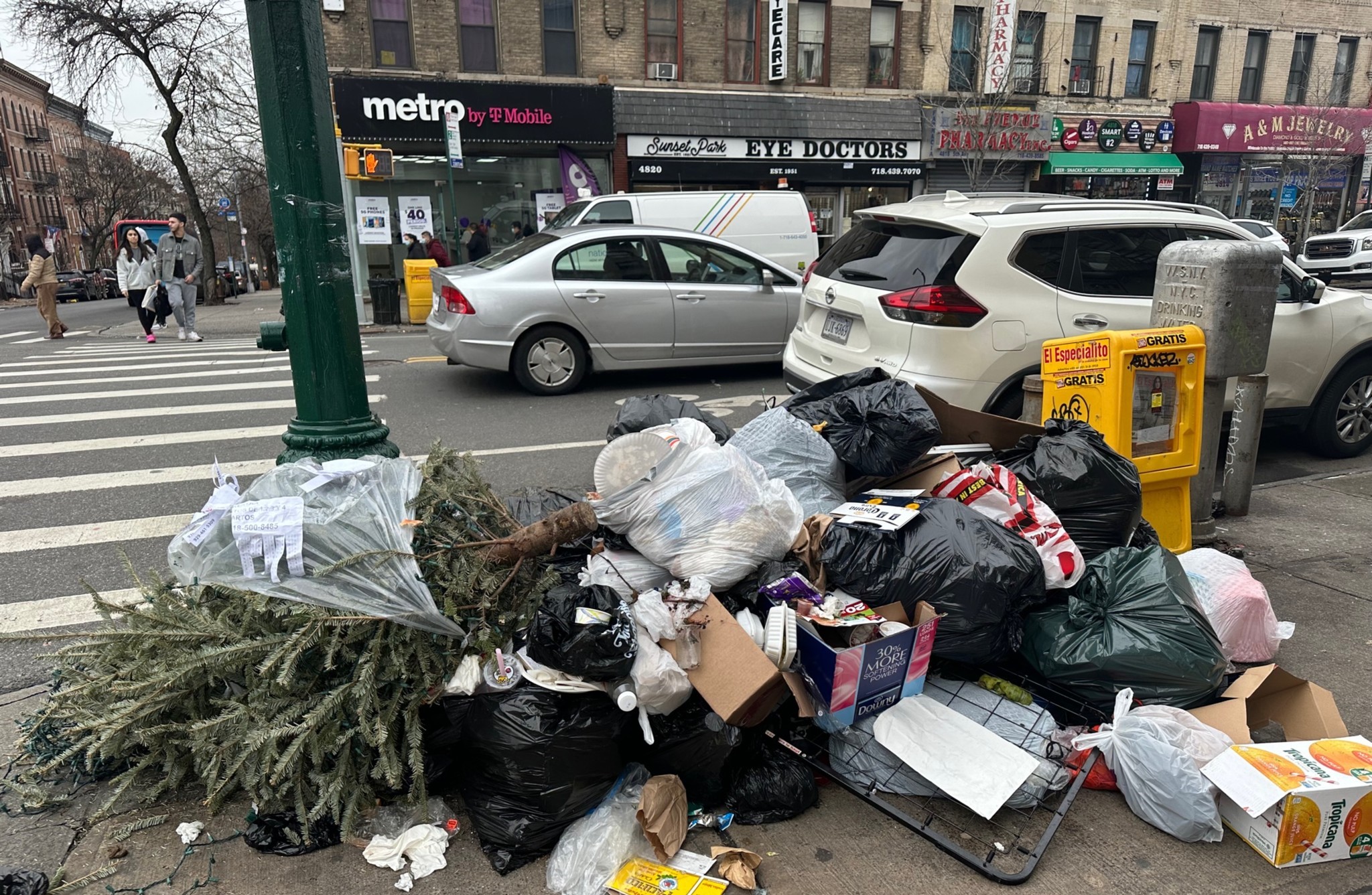 A pile of garbage, including trash bags and a discarded Christmas tree, is stacked on a sidewalk near a busy intersection. Cars and storefronts line the street.
