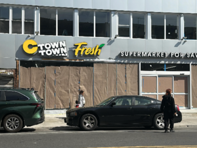 Two people stand on the sidewalk in front of a supermarket called "C Town Town Fresh." The store's windows are covered with brown paper, and two cars are parked along the street.