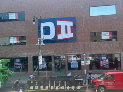 A brick building with a large "DII" sign and several "Grand Opening" posters in the windows. People work on the sidewalk near a red van and construction equipment.