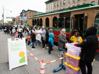 A long line of people, including children, wait at a bus stop next to a red and green trolley. A woman hands out items from plastic bins, and a sign in front reads "Sunset Park." The scene takes place on a city street.