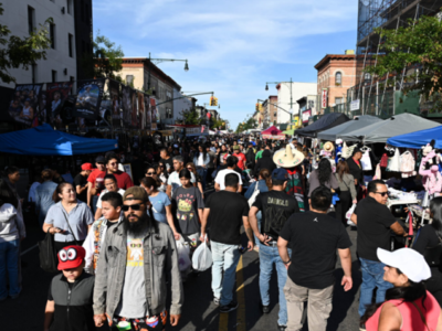 A large crowd of people walks along a city street lined with vendor booths and tents during a street fair or festival on a sunny day. Buildings and trees border the street.
