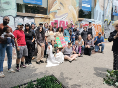 A diverse group of people poses together on a city sidewalk in front of a colorful mural and graffiti. Some hold a vibrant poster, while others stand or crouch, smiling for the camera. Greenery is visible in the foreground.